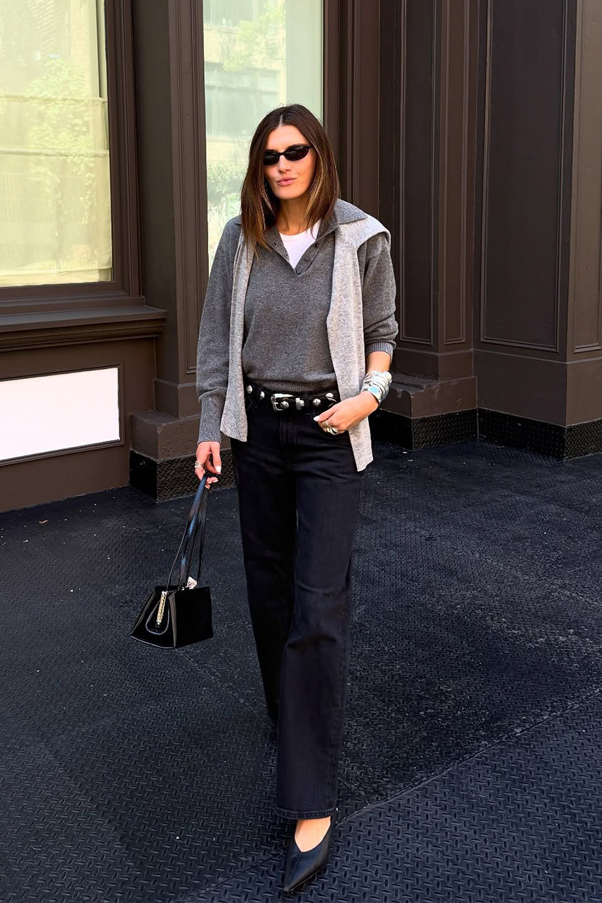 Layering staples for spring 2026 is shown in a street style photo with a woman standing outside while wearing a white t-shirt underneath a gray knit polo shirt styled with a light gray cardigan around the shoulders, black jeans, a black studded belt, black pointed heels, and a black handbag