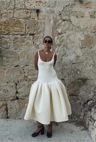 a photo of a woman's spring vacation outfit with a shell necklace, black sunglasses, and white drop waist dress and black pumps