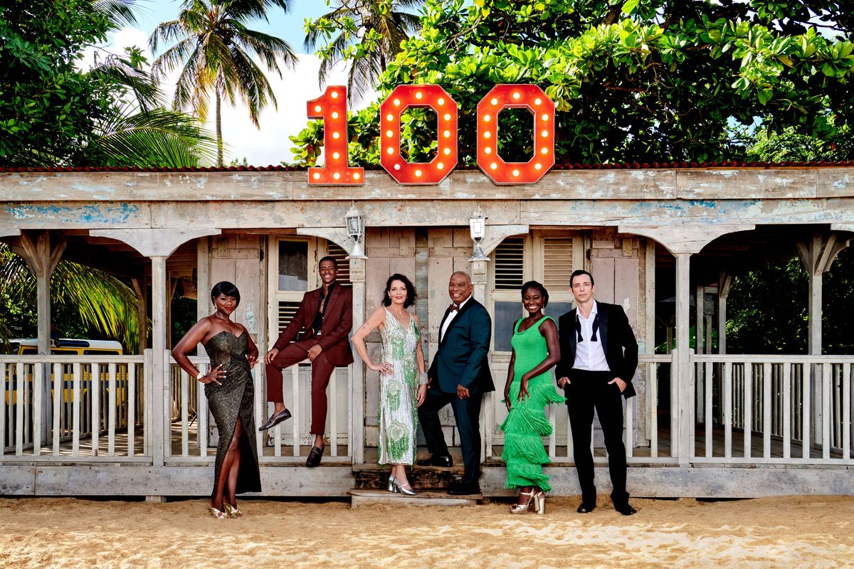 The cast of Death In Paradise pose in eveningwear outside of DI Neville Parker&#039;s beach hut, which has the number &quot;100&quot; displayed above it in lights. From left to right: Ginny Holder (Officer Darlene Curtis), Tahj Miles (Officer Marlon Pryce), Élizabeth Bourgine (Catherine Bordey), Don Warrington (Commissioner Selwyn Patterson), Shantol Jackson (DS Naomi Thomas) and Ralf Little (DI Neville Parker)