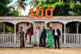 The cast of Death In Paradise pose in eveningwear outside of DI Neville Parker's beach hut, which has the number "100" displayed above it in lights. From left to right: Ginny Holder (Officer Darlene Curtis), Tahj Miles (Officer Marlon Pryce), Élizabeth Bourgine (Catherine Bordey), Don Warrington (Commissioner Selwyn Patterson), Shantol Jackson (DS Naomi Thomas) and Ralf Little (DI Neville Parker)