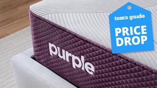 A closeup of the Purple logo at the foot of the Purple Restore Mattress on a bed frame, a Tom's Guide Price Drop deals badge in the top right corner