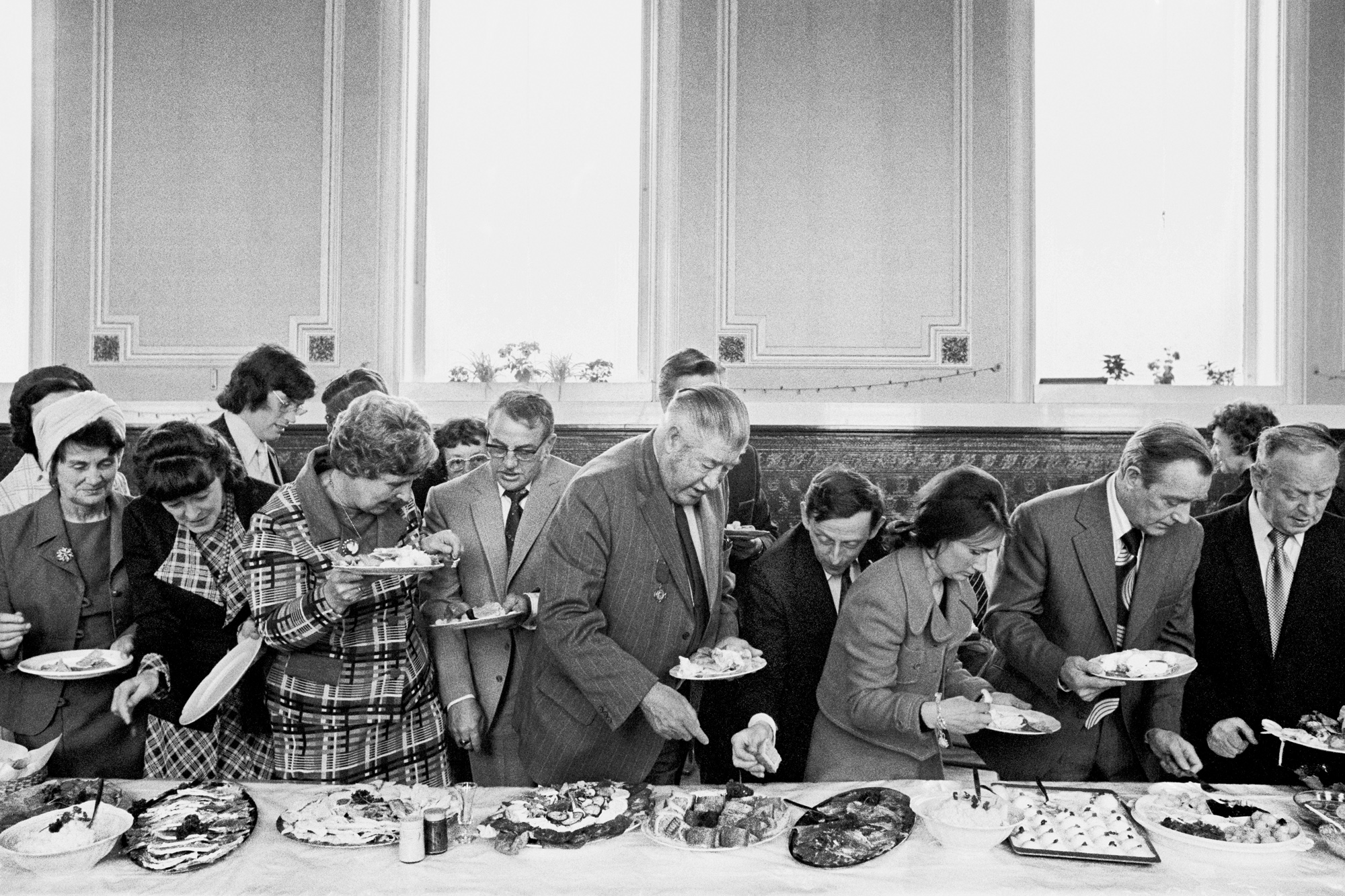 A black and white photo shows a line of formally dressed adults serving themselves food from a buffet table indoors, illuminated by large windows in the background.