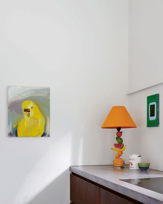 a corner of a kitchen with concrete benchtops, timbe cabinets, white walls, a table lamp with fruit details, and an artwork of a yellow budgie