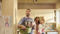 A dad looks up at the ceiling where water is dripping while Mom, holding their toddler, is on the phone in the background.