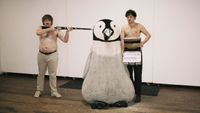 Getdown Services posing topless, with one of them pointing a rifle at the head of a giant plushie penguin