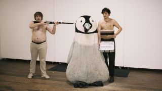 Getdown Services posing topless, with one of them pointing a rifle at the head of a giant plushie penguin