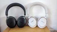 A white pair of Bose QC Ultra Headphones (2nd Gen) standing next to their black 1st Gen predecessors on a wooden surface in front of a white wall.