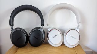 A white pair of Bose QC Ultra Headphones (2nd Gen) standing next to their black 1st Gen predecessors on a wooden surface in front of a white wall.