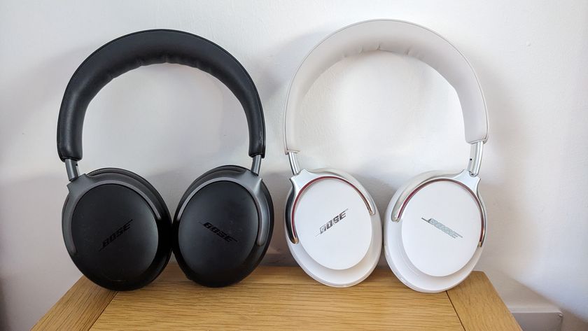 A white pair of Bose QC Ultra Headphones (2nd Gen) standing next to their black 1st Gen predecessors on a wooden surface in front of a white wall.