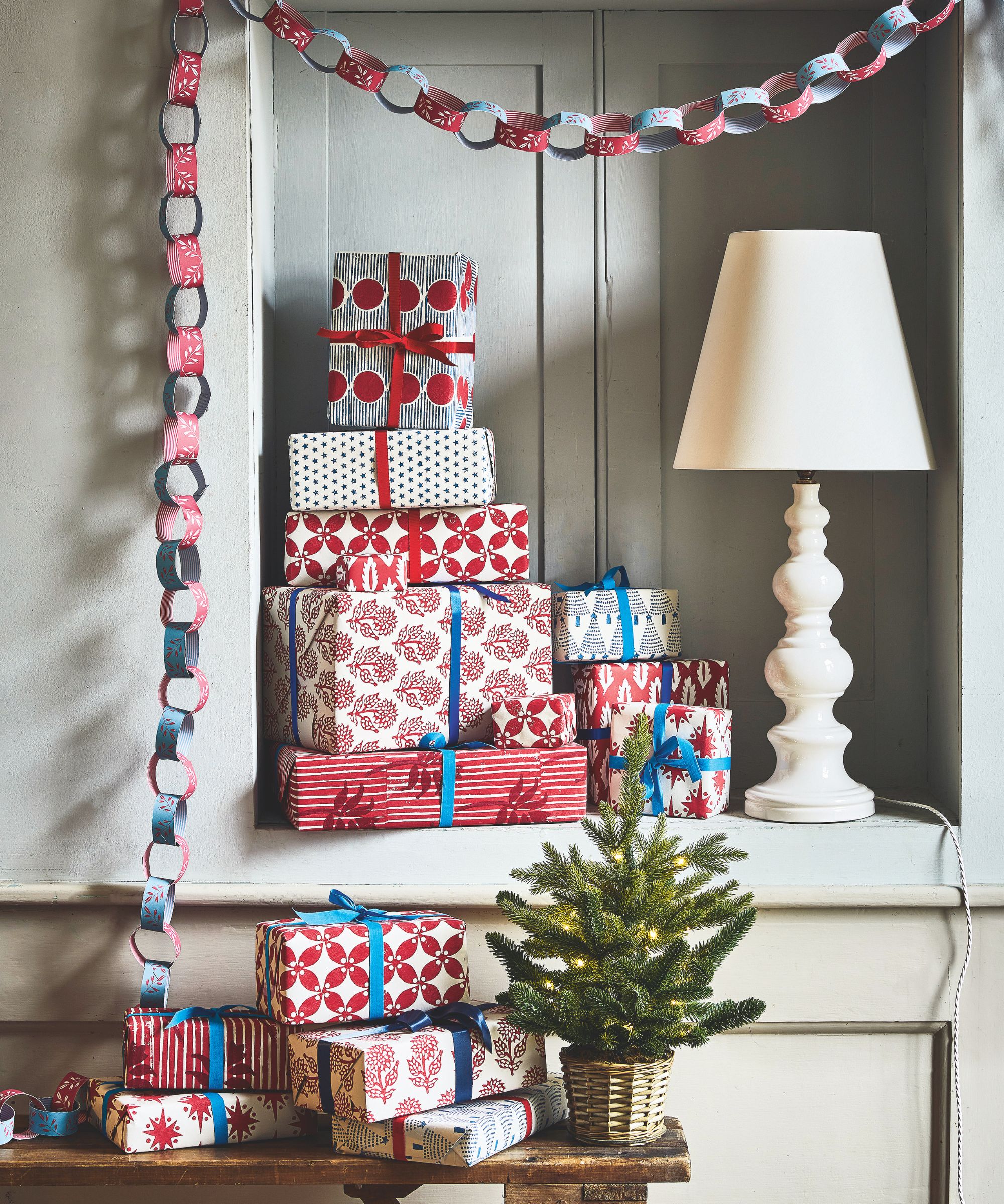 A pile of Christmas presents in patterned paper in an alcove beside a white lamp with a wooden table of presents in the foreground. Beside the presents is a small lit Christmas tree in a wicker pot, and hanging over the alcove are blue and red paper chains.