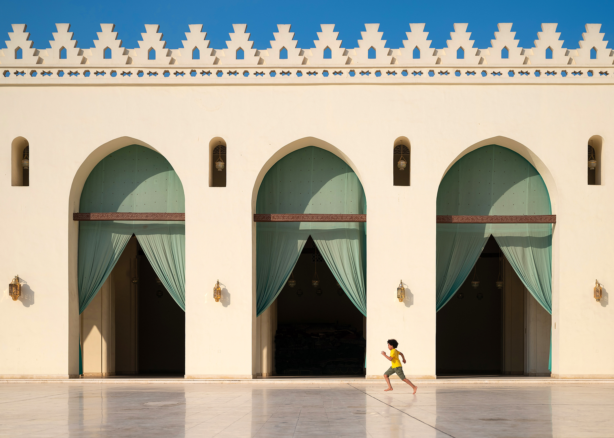 A young boy in a yellow shirt runs joyfully across a marble courtyard, framed by arched doorways with turquoise drapes