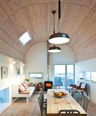 Large living-dining space with barrel vaulted ceiling, cladded in light wooden panels, large wooden dining table, black pendants hanging above, one skylight on ceiling, looking onto seating area with stove