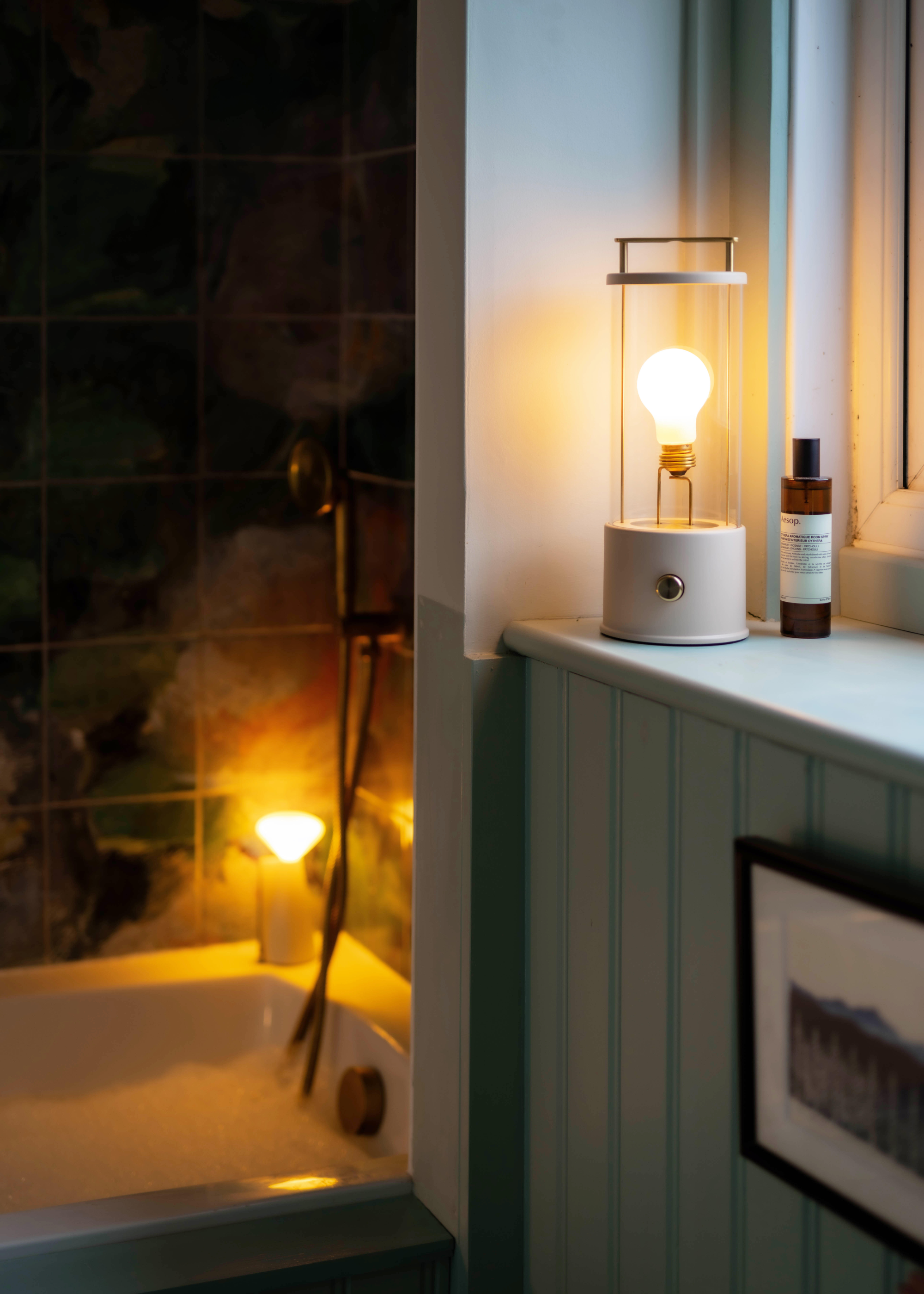 A portable lamp placed by a window sill next to a bottle. There is also a view of the bathtub with a lamp placed next to it.