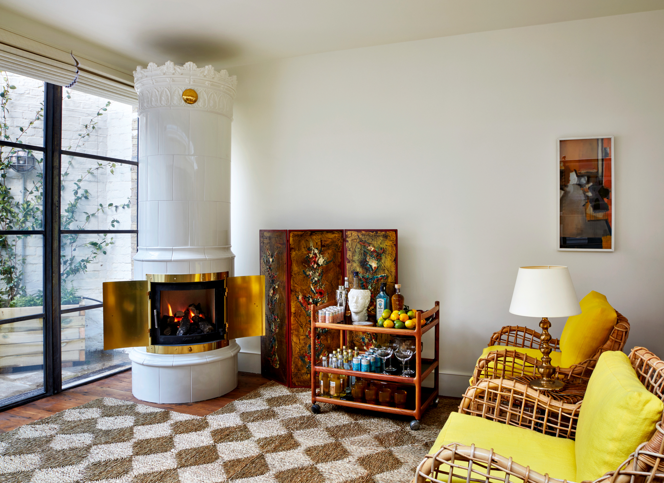 White kachelofen in a living room with yellow and cane chairs, wood bar cart and checkerboard rug