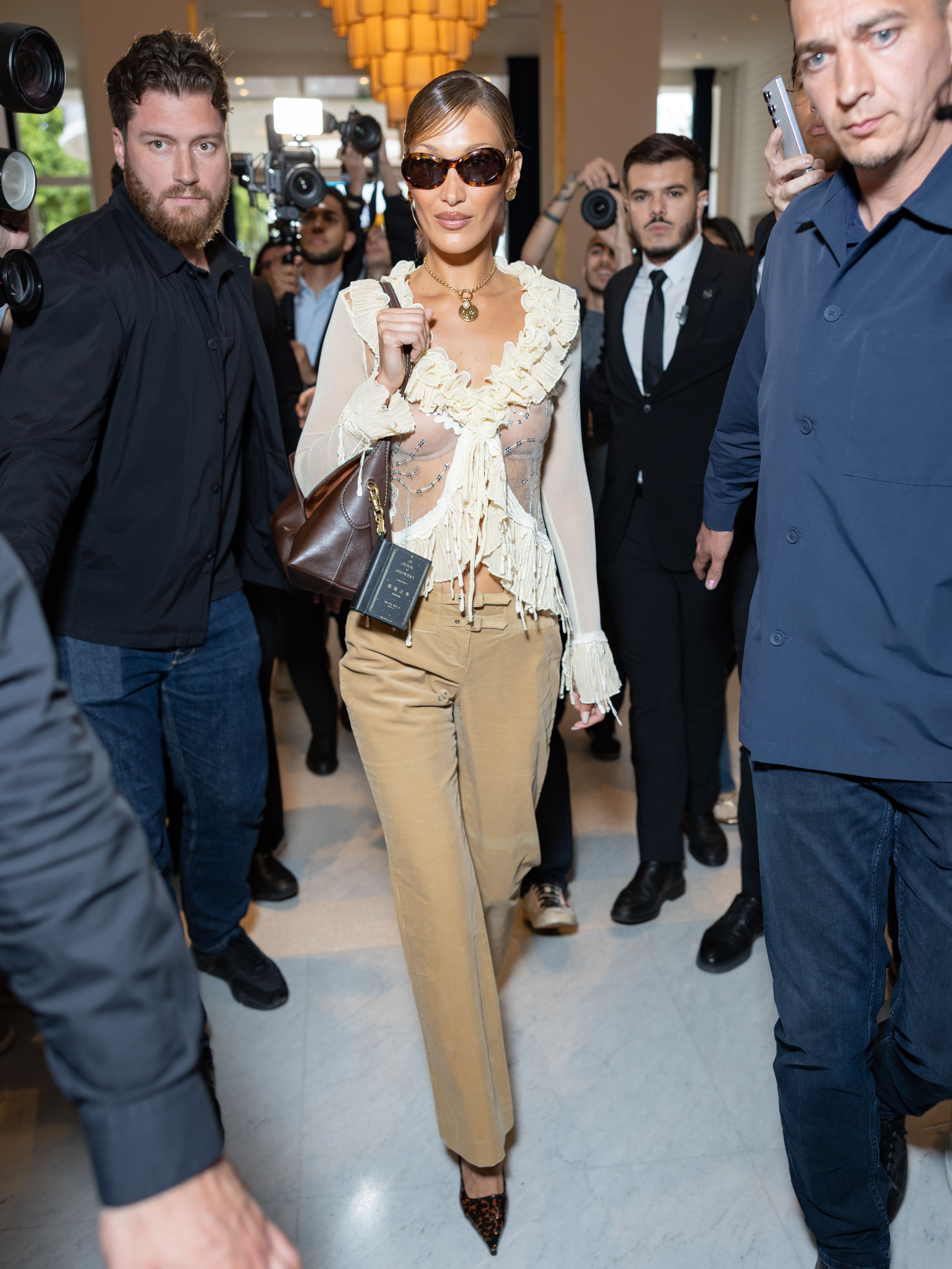 Bella Hadid wears a white ruffle blouse, beige pants, a Coach bag, and tortoiseshell heels.