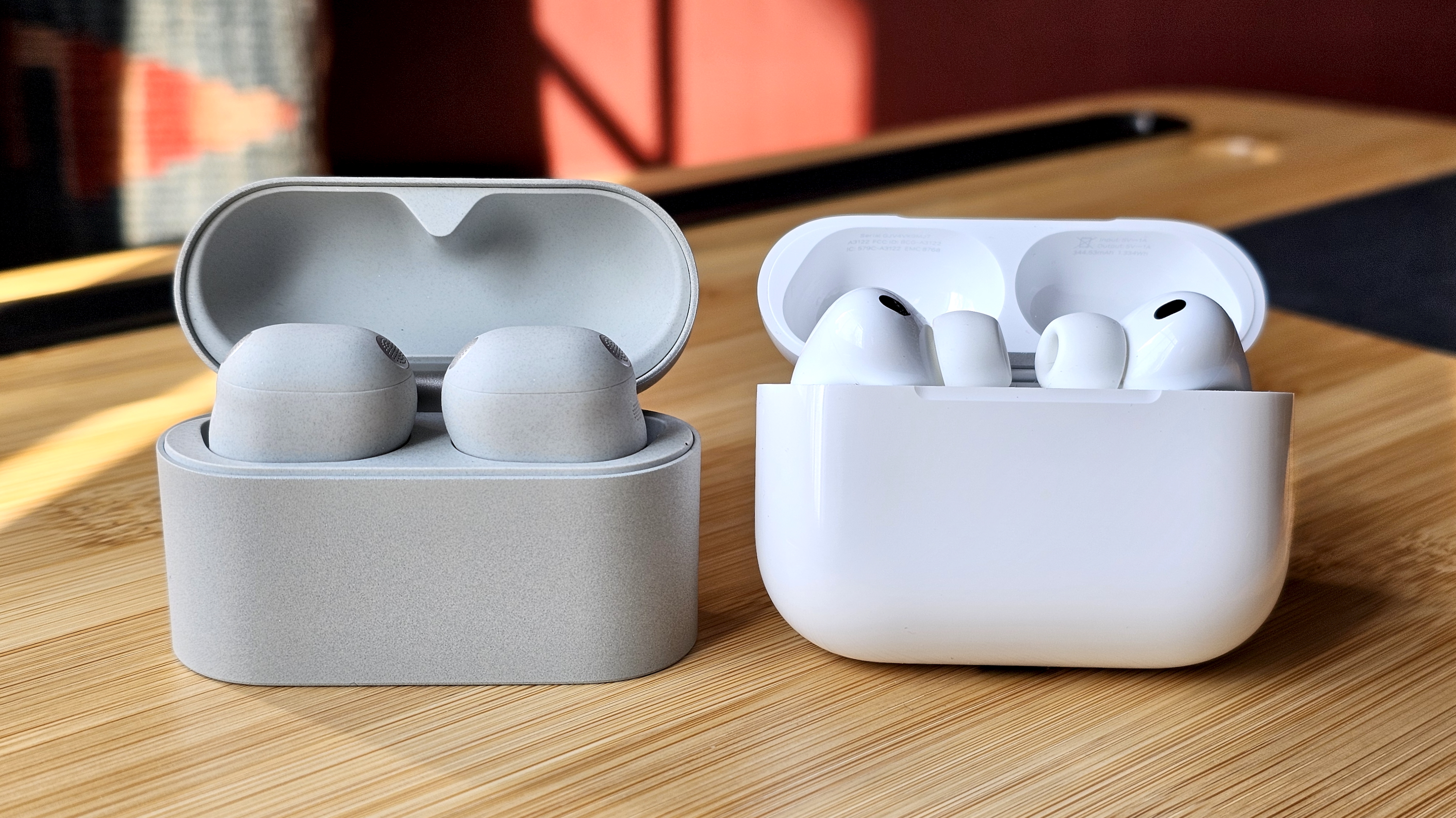 Sony WF-1000XM6 earbuds next to the Apple AirPods Pro 3, on a wooden table, in a versus-style format 