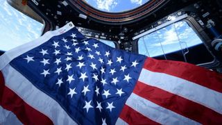 an american flag floats in a small circular room lined with windows. through the windows, earth can be seen far below