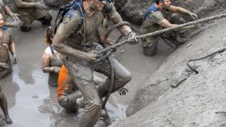 photo of a man coated in mud and holding a rope to climb up a wall in an obstacle course; others are pictured behind him, waiting in a pool of mud