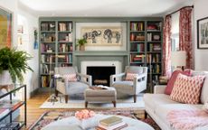 living room with green bookcase around fireplace, two armchairs, couch, rugs, floral drapes, pink cushions, footstool, plant