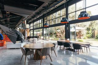 A naturally lit multifunctional space features Space Age design decor including orange and grey sculptural pendant lights, colossal sculptures, and blue and red accents while letting the surrounding nature come through its floor-to-ceiling loft-style windows.