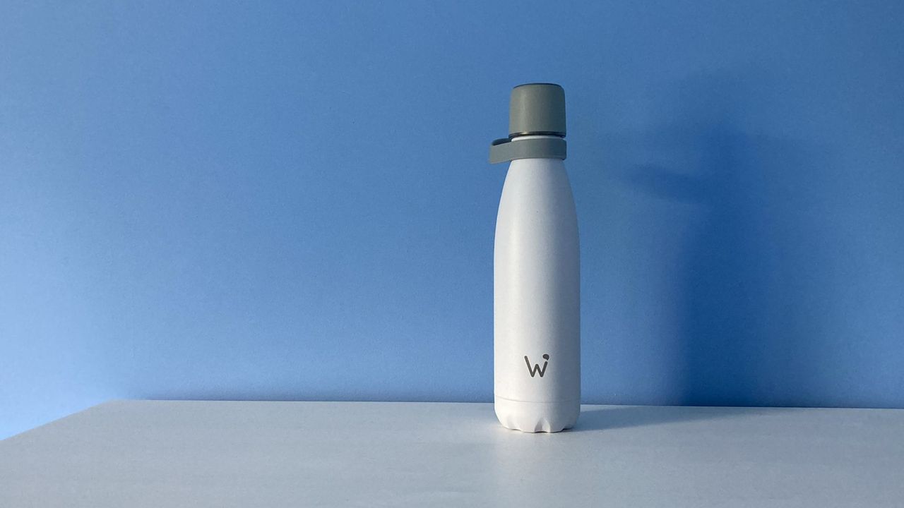 White water bottle with &quot;W&quot; logo in grey on the bottle and grey screw top, on white surface in front of a blue wall