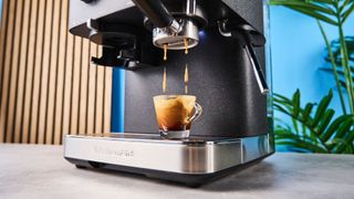 a black espresso machine by kitchenaid photographed against a blue background with silver chrome buttons and a pressure dial and steam wand