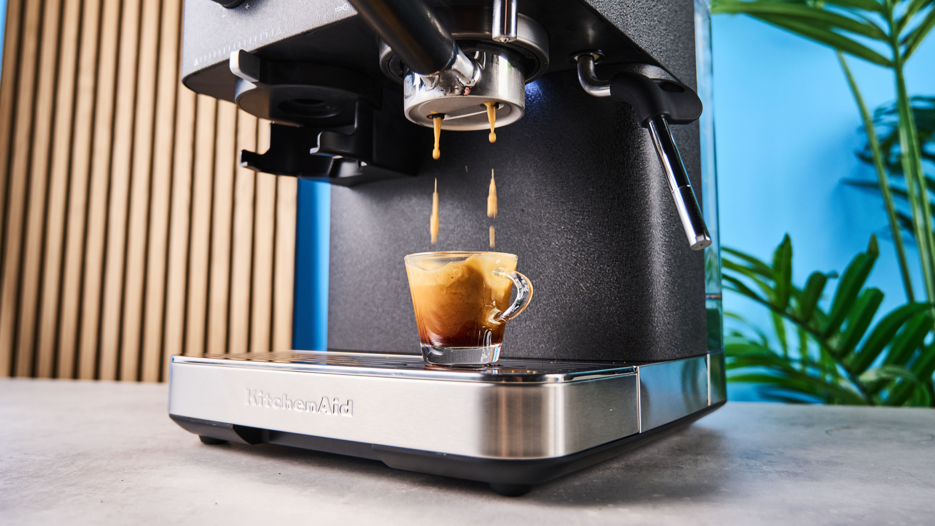 A black espresso machine from KitchenAid photographed against a blue background with silver chrome buttons, pressure regulator and steam wand.