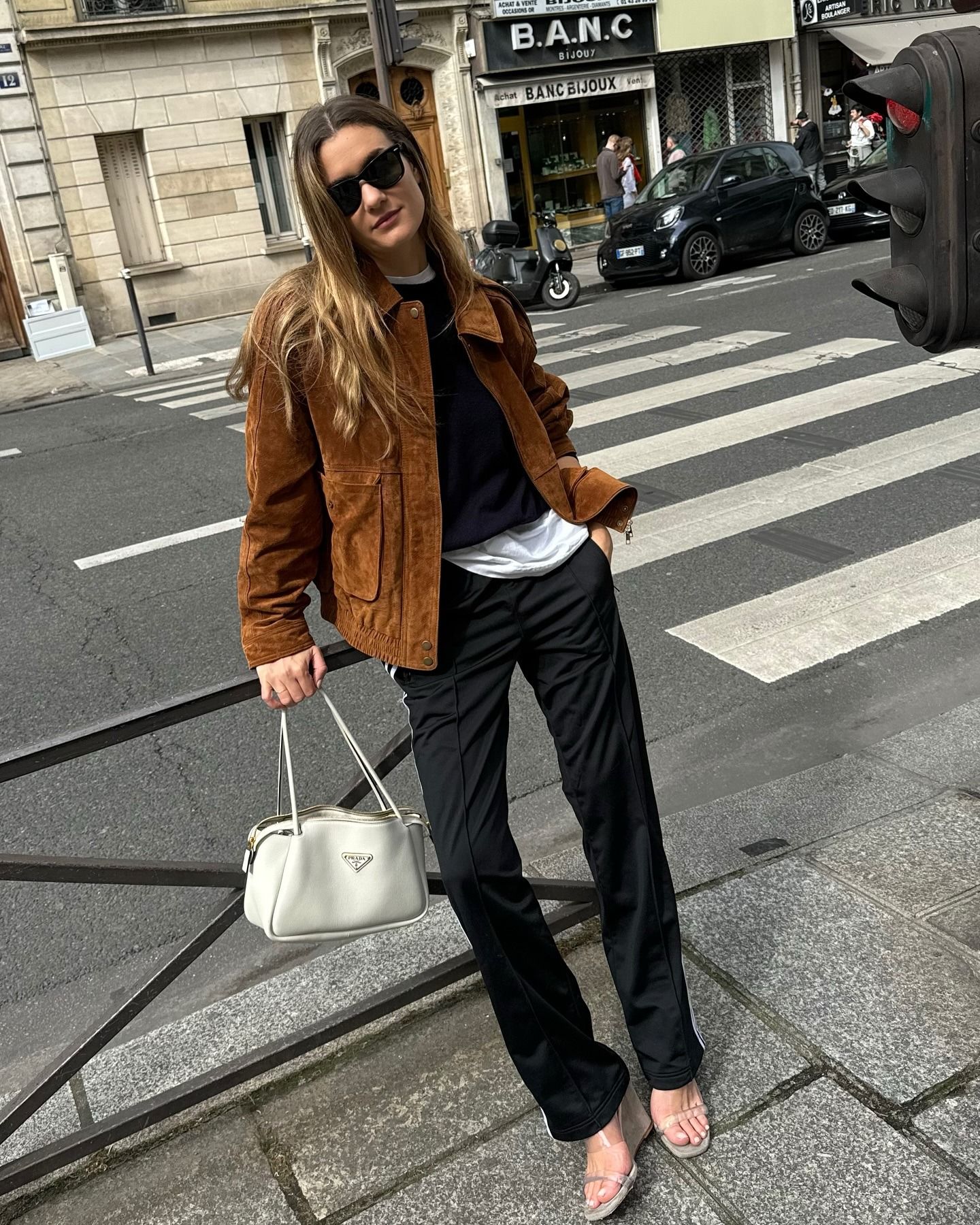 French influencer @annelauremais stands outside in Paris wear a brown suede jacket with black trousers and transparent heels.