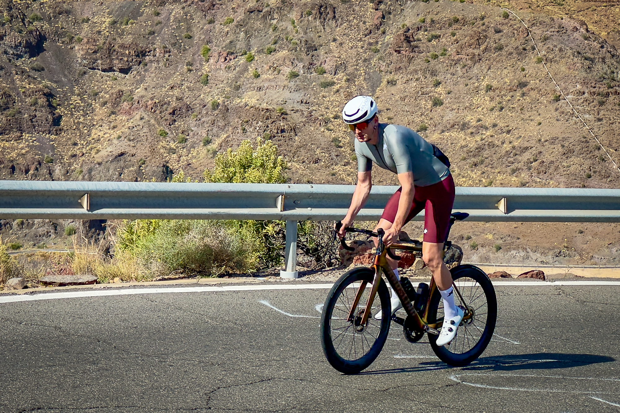 Man wearing a green jersey, red shorts and a white helmet riding an orange road bike up a sunny hill