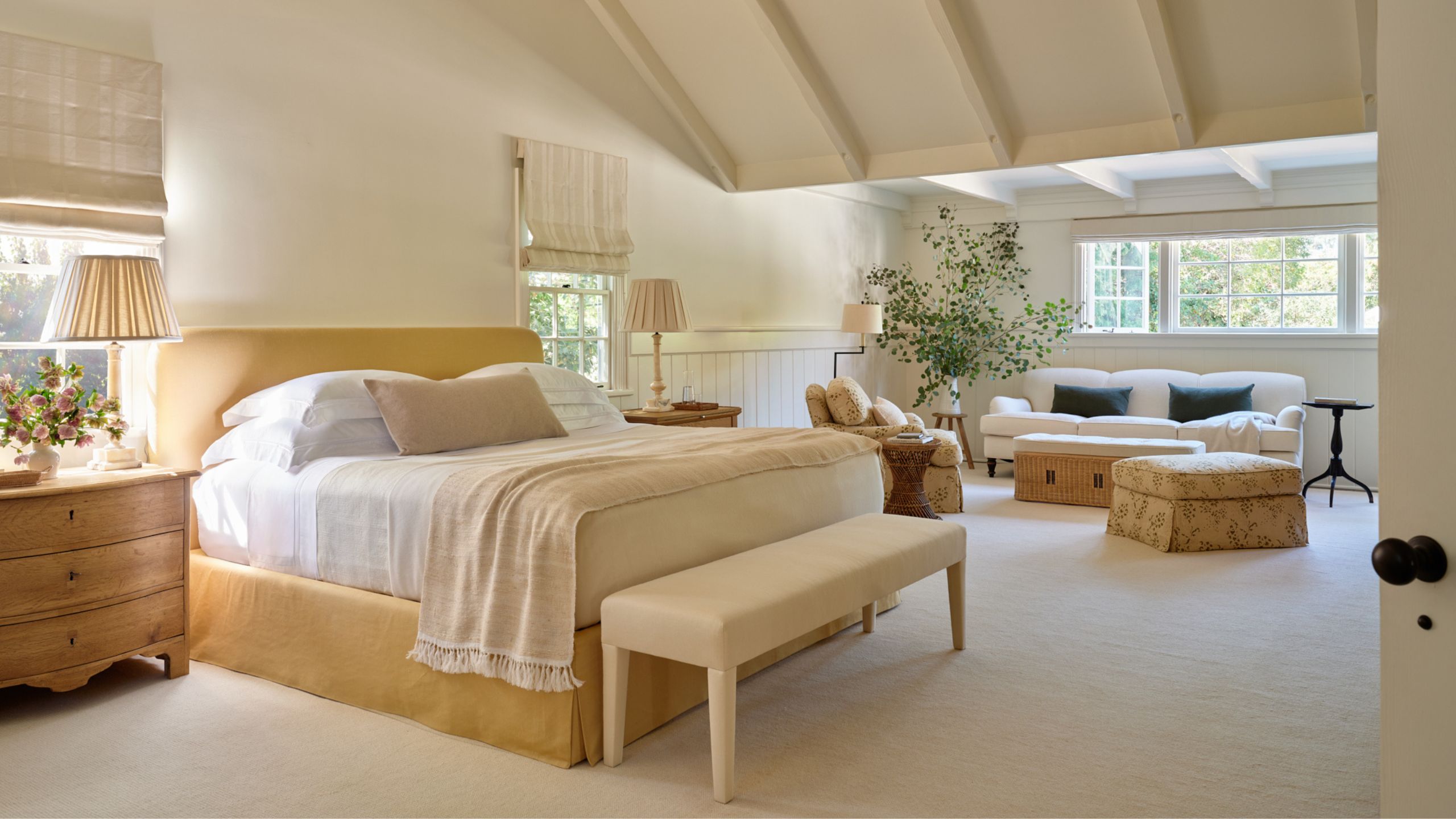 a large neutral primary suite with vaulted ceiling and a yellow upholstered bed, neutral linens, wooden nightstand and a cozy sitting area