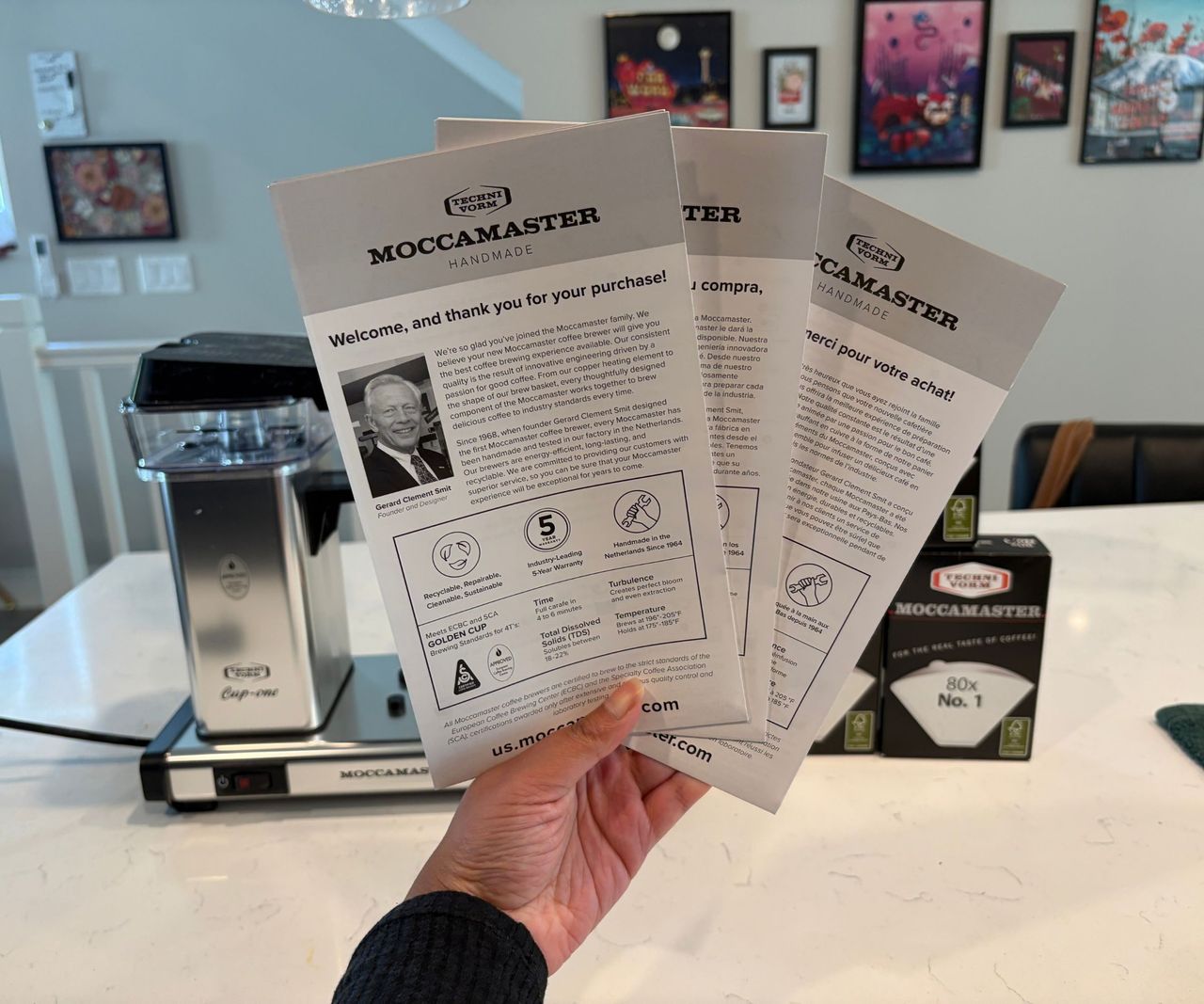 Someone holding the three instruction manuals for the Moccamaster cup-one coffee maker, with the coffee maker in the background.