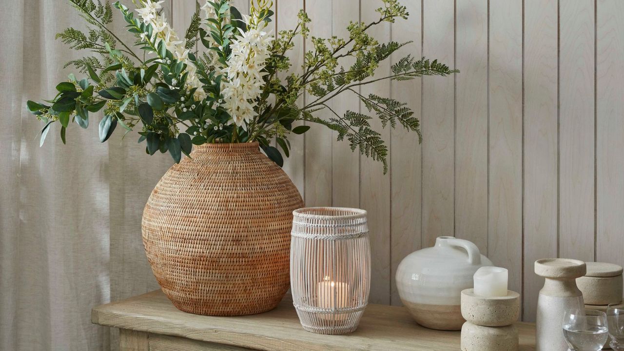 A woven wicker vase of hyacinth flowers and green stems next to a rattan candle holder with a lit pillar candle inside. On a wooden side table against a white painted shiplap wall. 