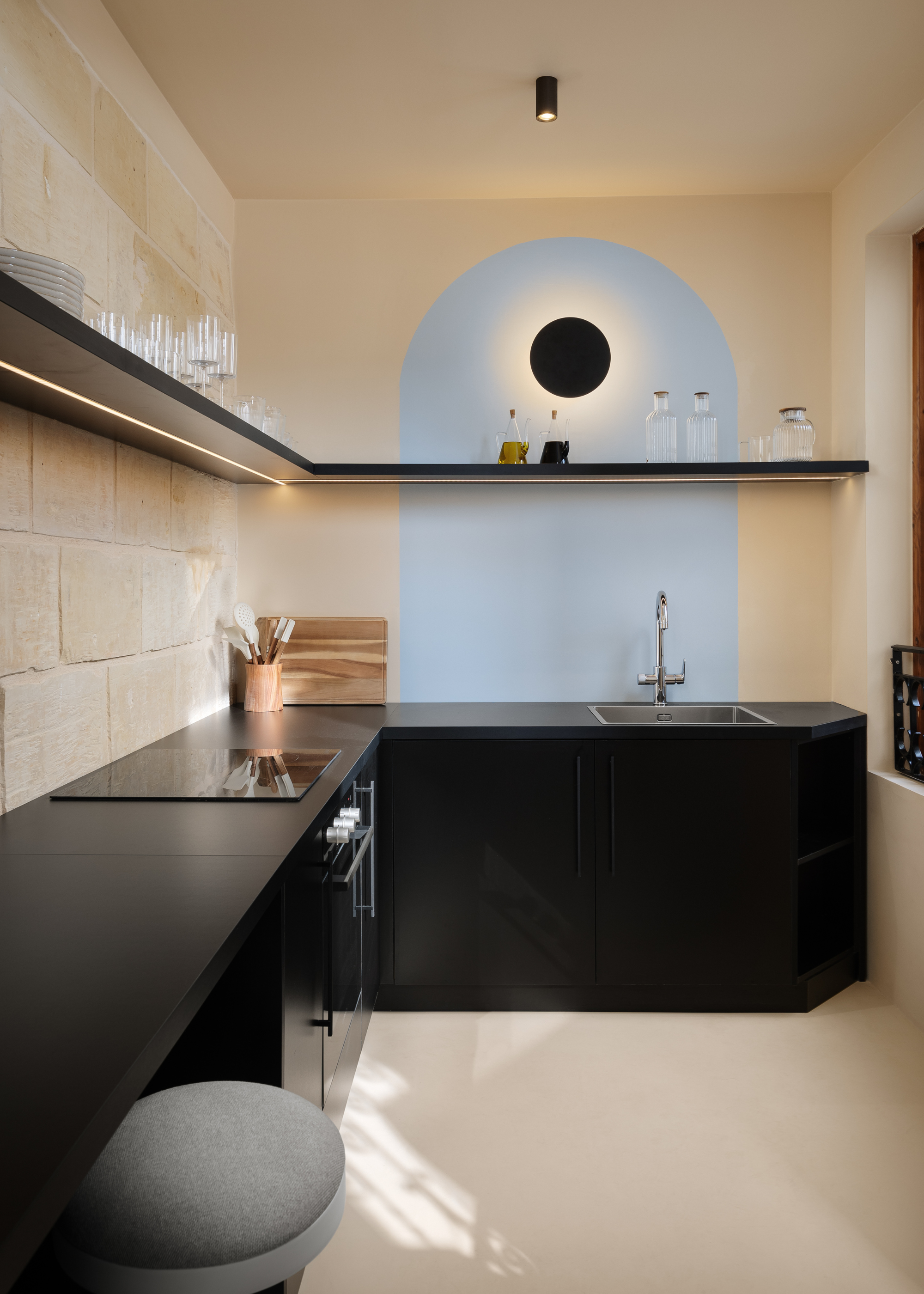 An off-white kitchen with a sky blue accent wall, black countertops, a gray stool, and floating shelves