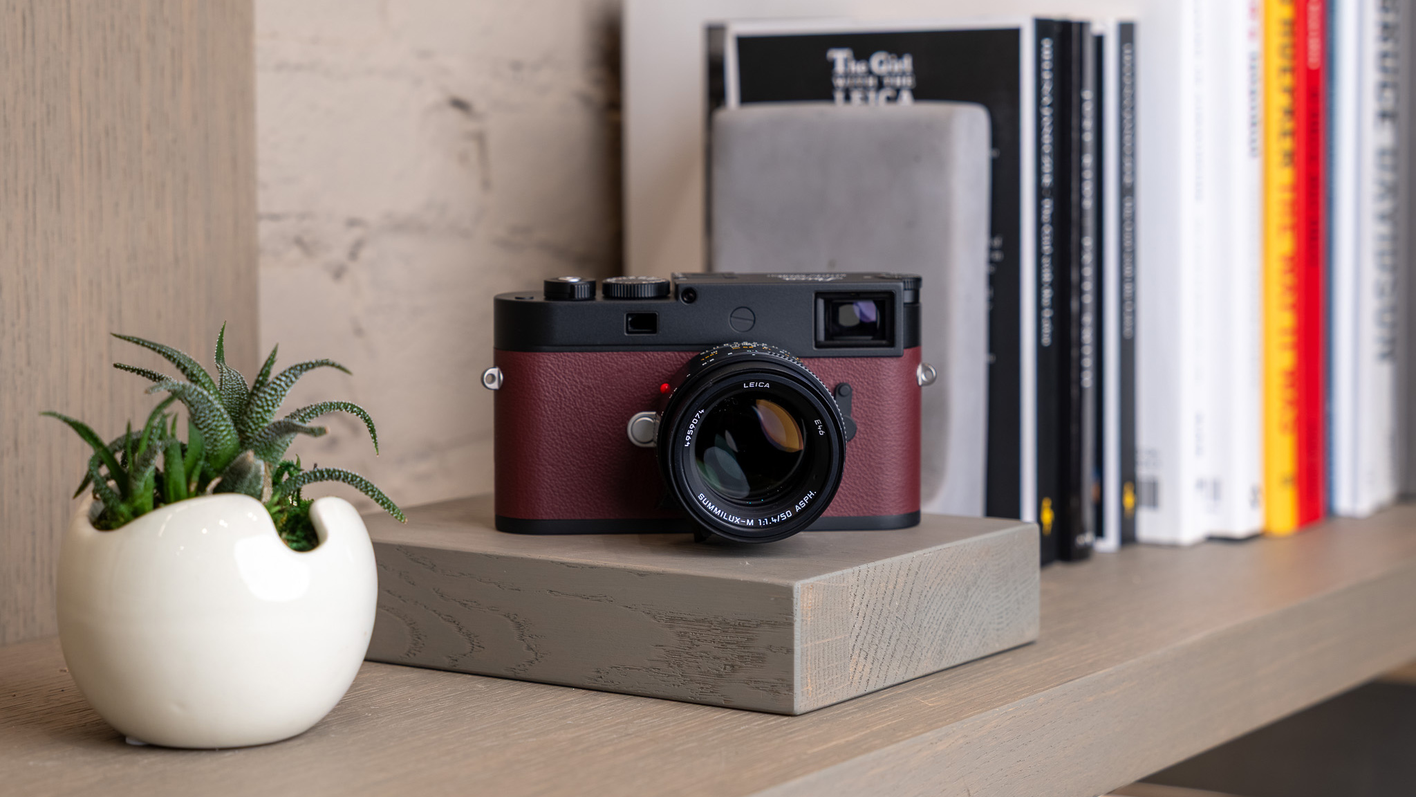 The Leica Chicago Edition 17 camera with lens in dark red, placed on a wooden shelf with a books and a small plant