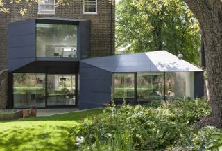 a very modern extension added on to a brick home, with the garden and bishes in the foreground