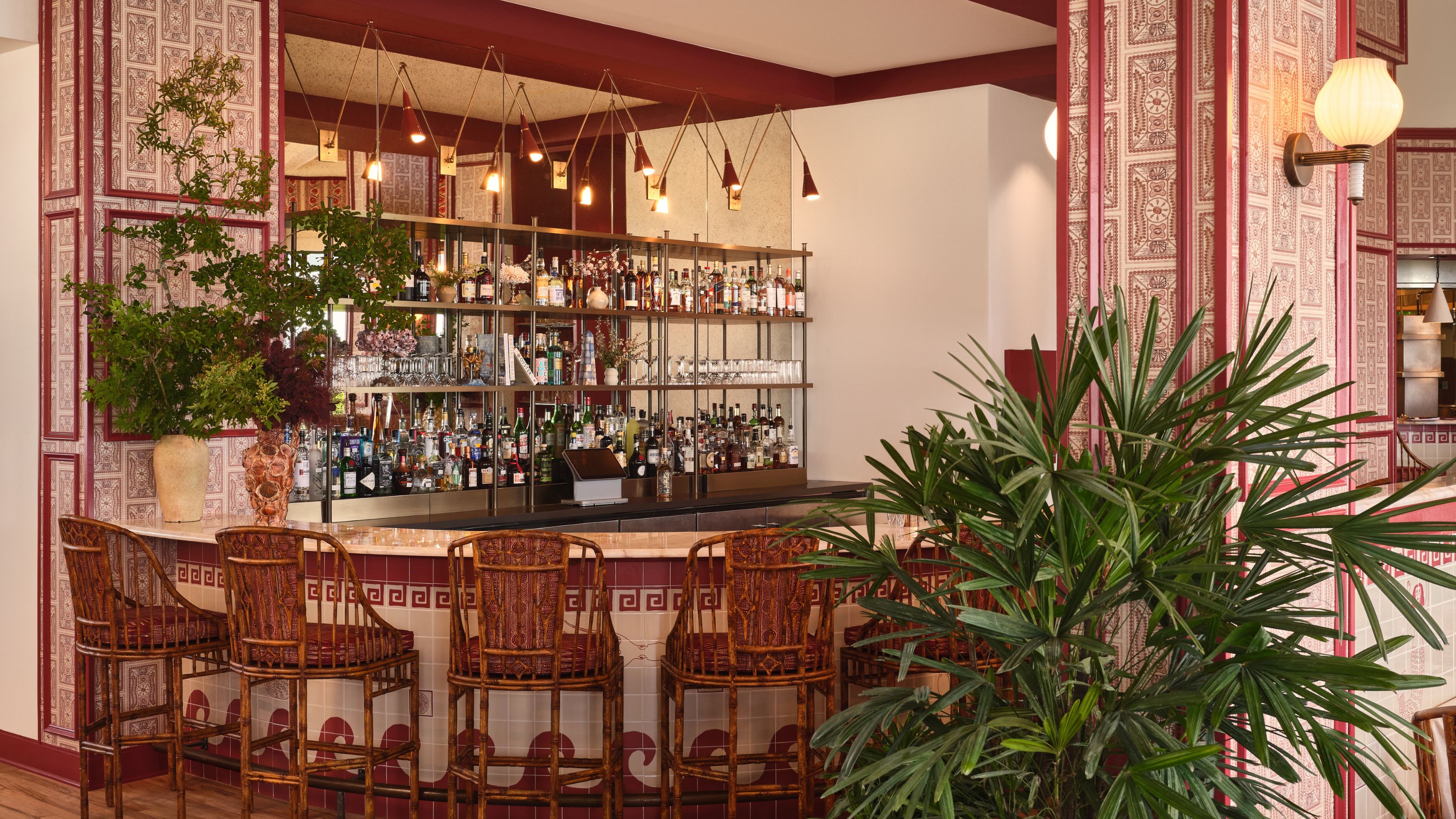 A retro-inspired bar area featuring a curved tile counter, woven bamboo barstools, elaborate hanging pendant lights, and patterned wallpaper columns.
