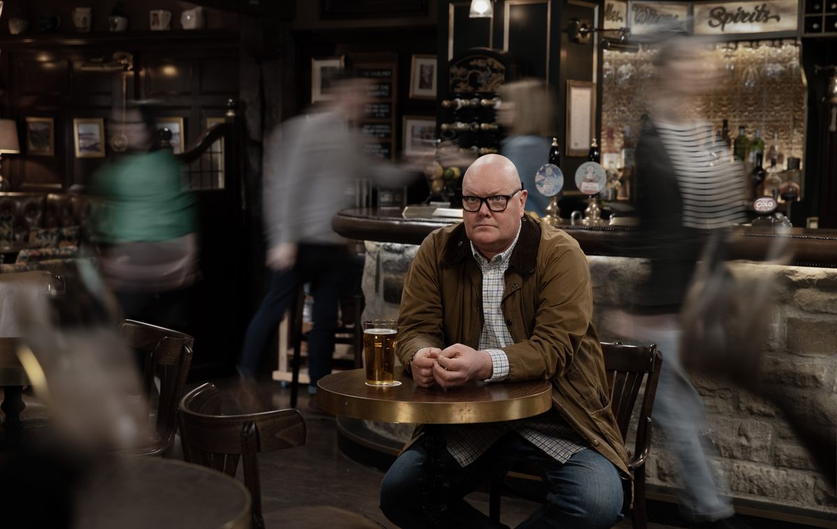 Emmerdale Paddy Kirk sitting in the Woolpack with blurry people around him. 