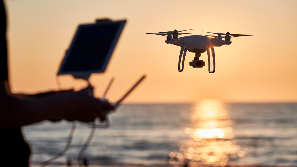 A person operates a drone against the light of a sunset on the ocean.