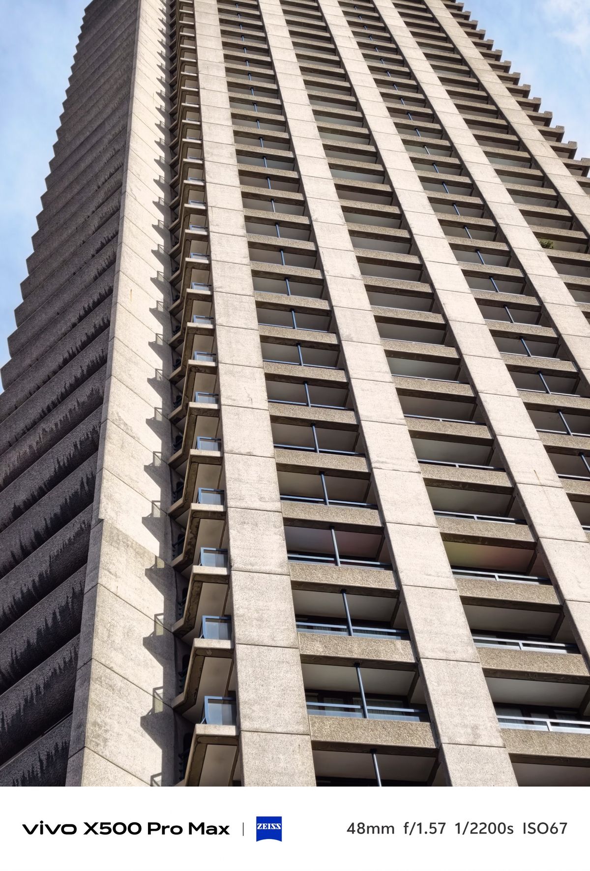 Low-angle view up a tall concrete residential tower with repeating balconies against a blue sky.