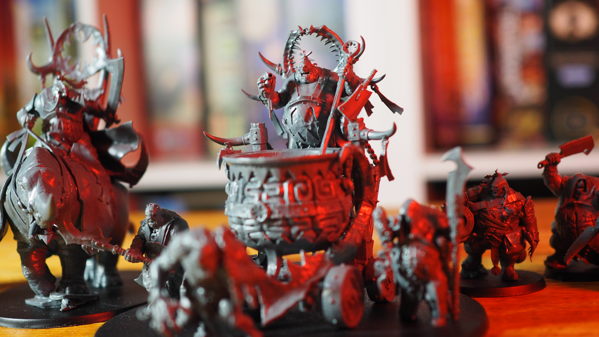 A collection of Ogor Mawtribes models on a wooden surface in front of a blurred shelf of board games