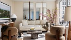 bright white living room with two mirrors on the wall behind a white sofa, console with lamp and books and art hung above, two tan armchairs on either side of a marble coffee table
