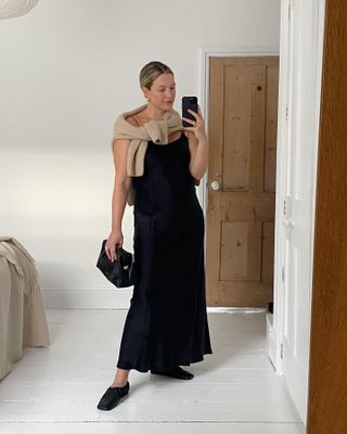 British fashion influencer Alexis Foreman poses for a mirror selfie wearing a black slip dress, a tan sweater tied around her shoulders, a black clutch bag, and black glove flats