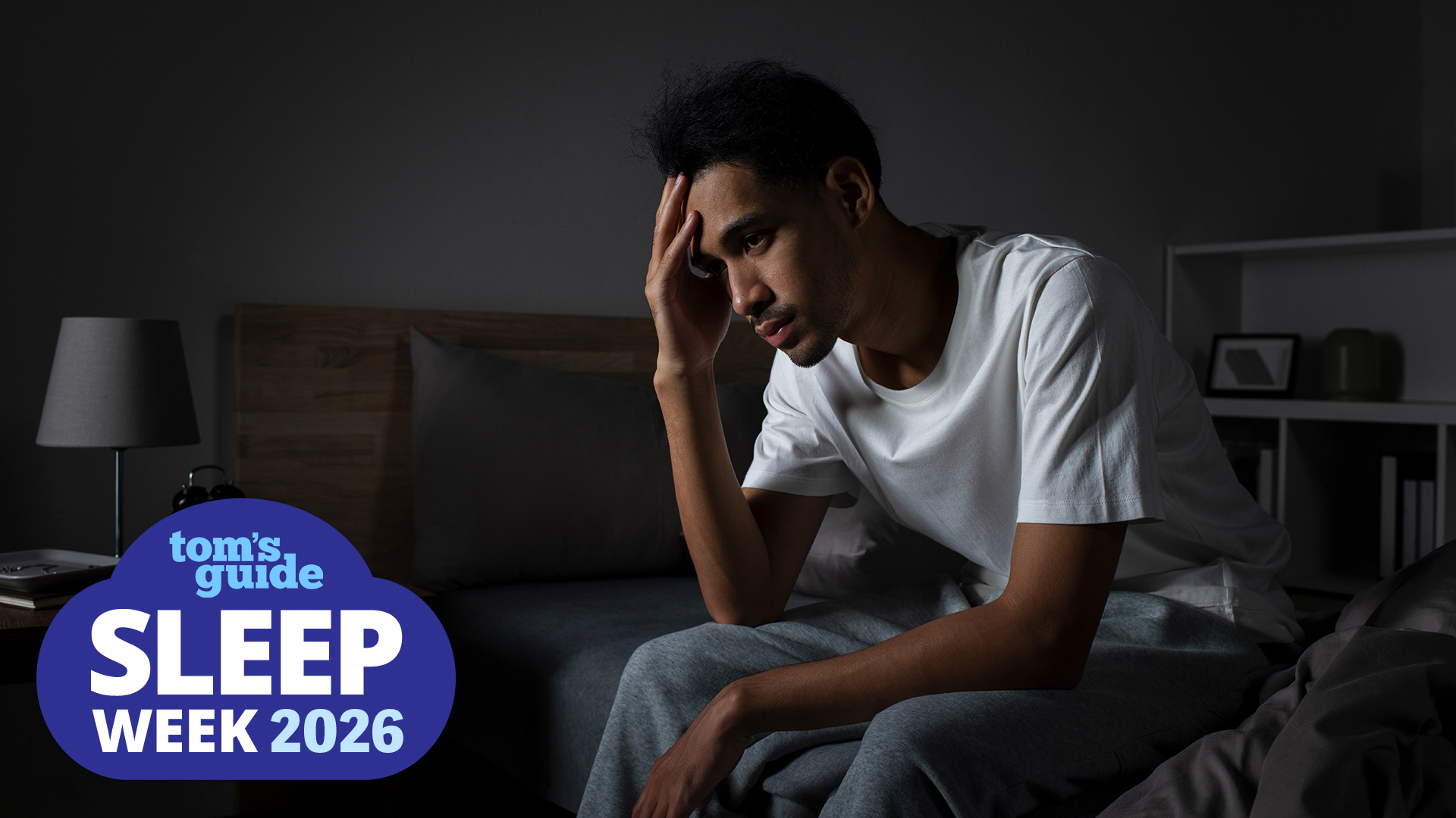 A man sits on the edge of his bed in a dark room with his head in his hands, unable to sleep. In the bottom left corner is a Tom's Guide sleep week deals graphic