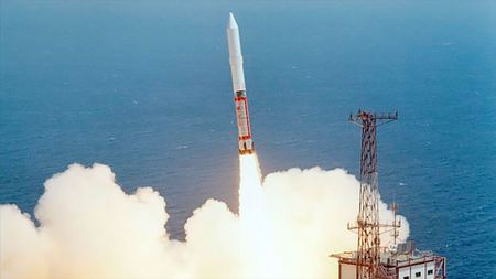 a red and white rocket lifts off at an angle as viewed from above, setting it against the blue of the ocean beyond the launch pad.