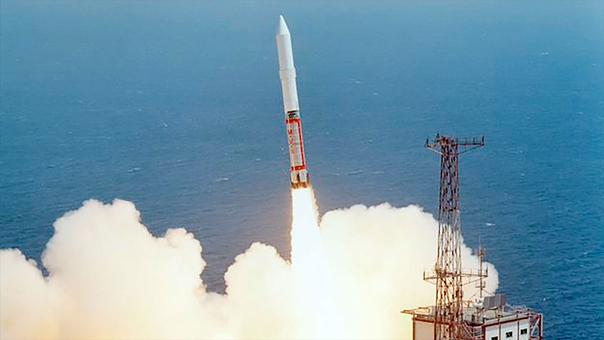 a red and white rocket lifts off at an angle as viewed from above, setting it against the blue of the ocean beyond the launch pad.
