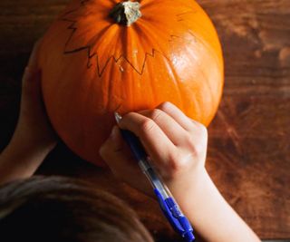 Two hands drawing an outline on a large orange pumpkin