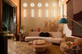 Image of an eclectic seating room. There is a curved, patterned sofa at the back of the room, with a round wooden coffee table and a brown furry accent chair in front of the sofa. The walls have unique, geometric lighting and room divider screen. There is a cutout rug hanging on the wall.