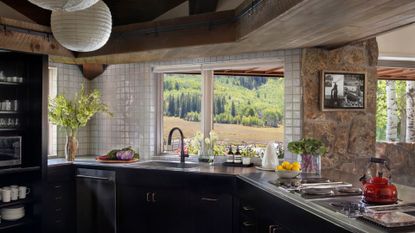 A bright kitchen with a tiled backsplash, a steel countertop, windows by the sink, vases of flowers, fresh produce, a red kettle, and a pair of hanging rice paper lamps