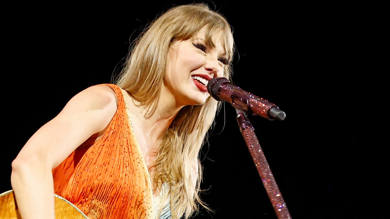 Taylor Swift wearing an orange fringed dress singing into a red microphone and holding a guitar on stage