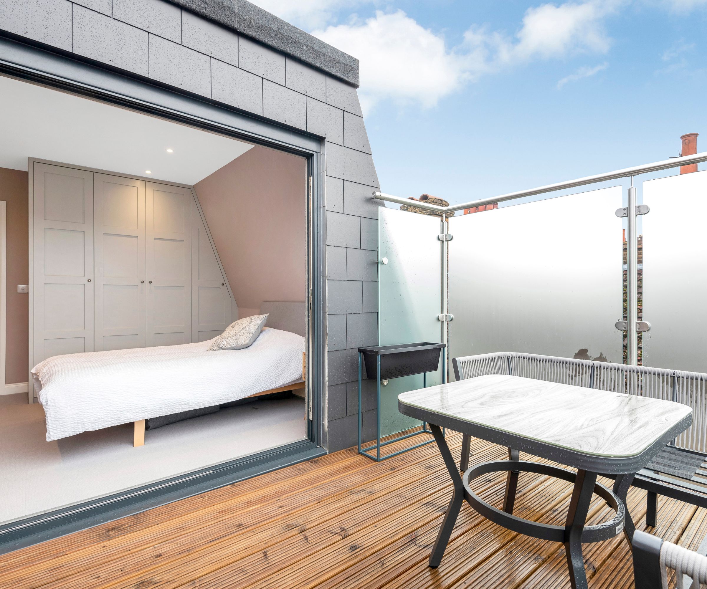 Modern loft bedroom with folding glass doors opening onto a compact balcony with decking, a bistro table and frosted glass balustrade.